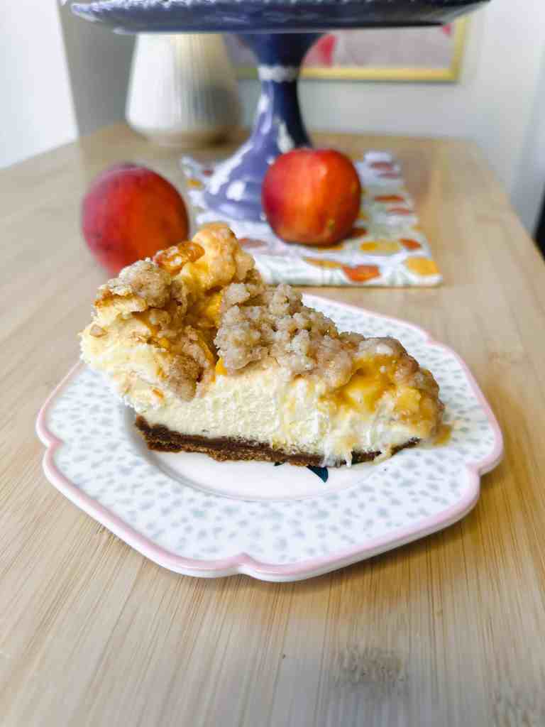 A close-up view of a slice of Peach Crumble Cheesecake showing the creamy cheesecake layer, juicy peach pieces, and a crunchy crumble topping on a Biscoff cookie crust.