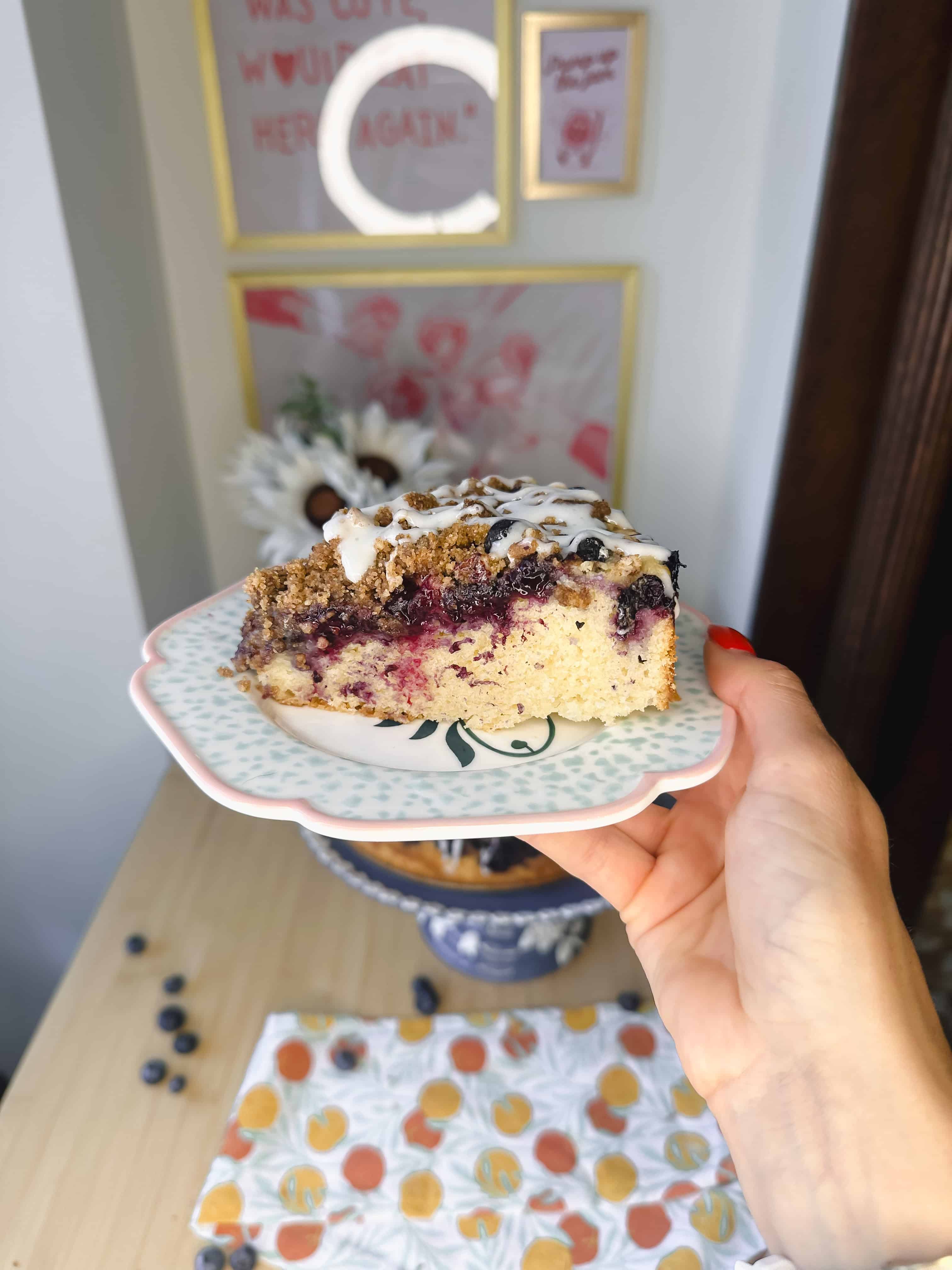A slice of blueberry crumble cake on a plate, showing layers of moist cake, juicy blueberries, crunchy crumble, and a drizzle of cream cheese glaze.